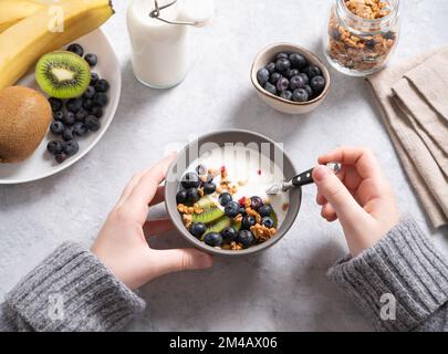 mani che tengono una ciotola con una sana colazione di yogurt, muesli e frutta fresca kiwi, banana e mirtillo su sfondo blu. Vista dall'alto. Foto Stock