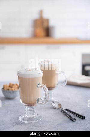 Due bicchieri di fragrante cappuccino con cannella su un tavolo blu sullo sfondo di una cucina bianca al mattino presto. Colazione Conc Foto Stock