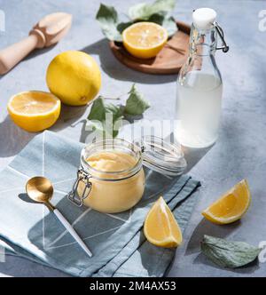 Deliziosa cagliata di limone fatta in casa in un vasetto di vetro con fette di limone e succo di bottiglia su sfondo blu. Colazione concettuale con ombre mattutine Foto Stock