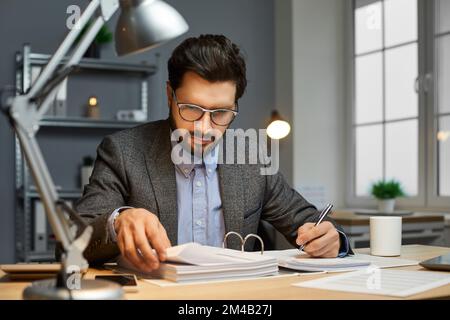 Contabile maschile seduto alla scrivania di un ufficio, facendo documenti e leggendo documenti Foto Stock