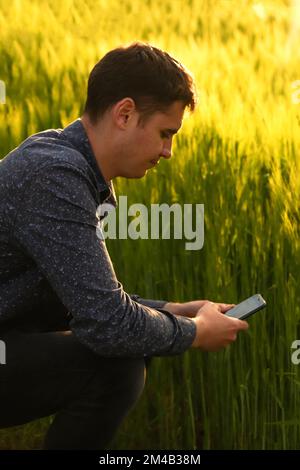 Sfocare l'uomo con il telefono al tramonto. Giovane agricoltore che lavora in un campo di grano, ispezionando e sintonizzando l'impianto sprinkler a perno del centro di irrigazione sullo smartphone. Foto Stock