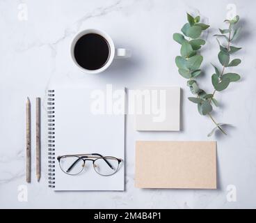 Piatto con fiore di eucalipto, tazza di caffè, matita, bicchieri, taccuino artigianale con pagine bianche su un tavolo di marmo. Desktop floreale da donna. mor Foto Stock