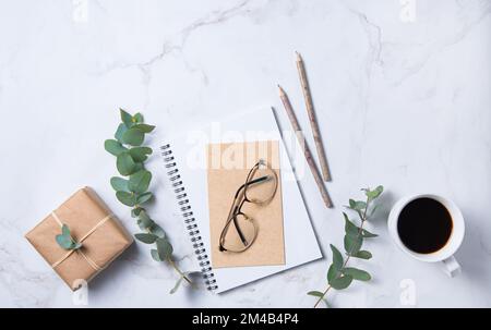 Piatto con fiore di eucalipto, tazza di caffè, bicchieri, matite, taccuino artigianale e scatola gigt su un tavolo di marmo. Desktop floreale da donna. mattina Foto Stock