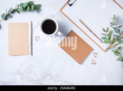 Piatto con fiore di eucalipto, tazza di caffè, matita, taccuino artigianale con pagine bianche su un tavolo di marmo. Desktop floreale da donna. mattina concep Foto Stock