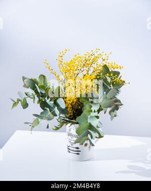 un bouquet primaverile con fiori mimosa gialli e rami di eucalipto si erge su una tavola bianca. Concetto del 8 marzo, giorno della donna felice. Spazio di copia Foto Stock