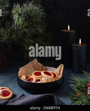 Biscotti fatti in casa con marmellata di frutta in una ciotola nera su un tavolo blu scuro con candele, tovagliolo e abete. Immagine scura e di umore Foto Stock