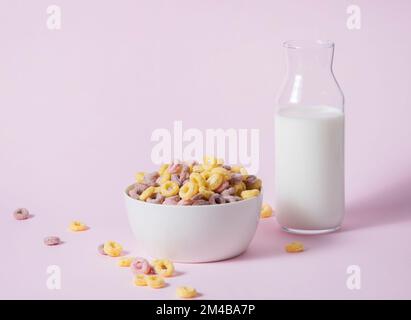 dolci cornflakes colorati anelli in un recipiente bianco con una bottiglia di latte su uno sfondo rosa. Vista frontale e immagine dello spazio di copia Foto Stock