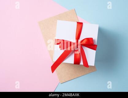 Piatto laico di celebrazione. Scatola regalo bianca con nastro rosso e busta artigianale su sfondo rosa pastello e blu. Concetto di San Valentino, Madre Foto Stock