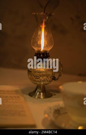 foto una tazza retrò vuota e un piattino si trova accanto ad un vecchio libro aperto e ad una lampada al cherosene che brucia Foto Stock