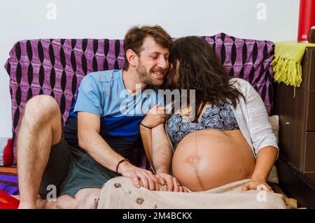 Donna adulta incinta che parla segretamente con suo marito un uomo adulta caucasico argentino sorridente, sono seduti sul divano all'interno della casa Chattin Foto Stock