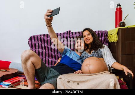 Coppia latina, uomo adulta caucasico con sua moglie, donna adulta brasiliana incinta, sta scattando una foto selfie con il cellulare, insieme seduto sul sof Foto Stock