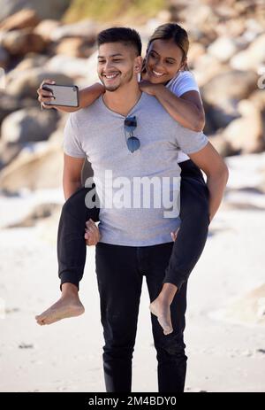 Coppia, piggyback e selfie sulla spiaggia per divertirsi e legarsi con amore e affetto. Uomo, donna e data oceano mentre si scatta una foto mobile sul Foto Stock