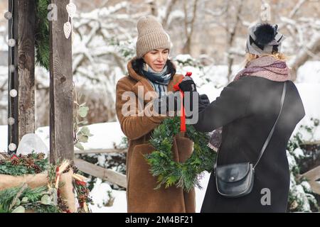 Bella coppia di ragazze in abiti caldi che comprano bella ghirlanda di Natale o corona in un mercato di strada in inverno con neve e fumo da cibo caldo Foto Stock