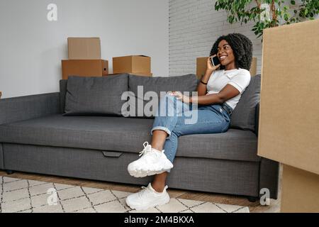 Giovane donna africana con smartphone e scatole che si sposta in una nuova casa Foto Stock