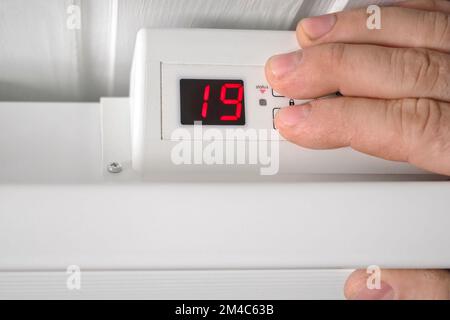 Uomo che abbassa la temperatura sul riscaldatore elettrico. Primo piano. Persona che regola il termostato sul radiatore per risparmiare energia a causa del costo del riscaldamento aumento del prezzo. ENER Foto Stock