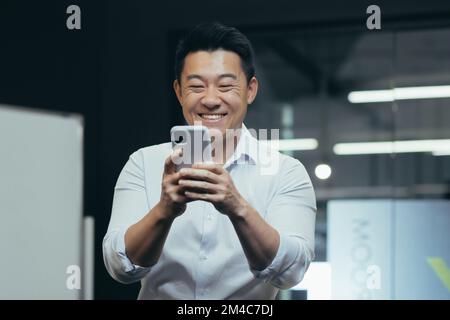 Un giovane uomo asiatico sorridente è in piedi in ufficio e utilizzando il telefono. Gioca a giochi online, digita messaggi, legge buone notizie. Foto Stock