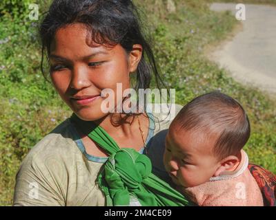 Raga, Arunachal Pradesh, India - 11 20 2013 : Ritratto all'aperto di bella giovane donna sorridente della tribù di Nyishi con il trasporto del suo bambino Foto Stock