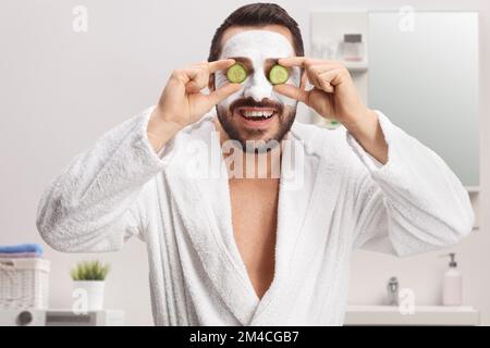 Uomo in un accappatoio con una maschera viso che tiene cetrioli sugli occhi in un bagno a casa Foto Stock