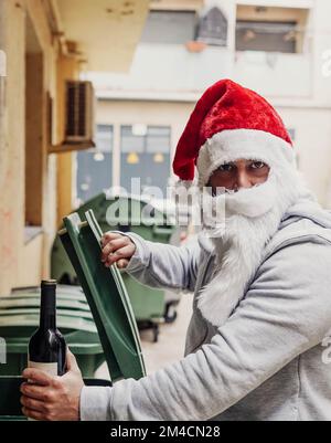 Babbo Natale senza tetto bere vino e rummaging attraverso bidoni della spazzatura Foto Stock