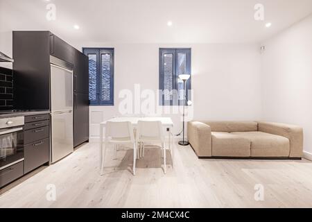 Soggiorno con cucina aperta di armadi neri con piastrelle abbinate, balcone tavolo da pranzo con sedie e due finestre con cornici dipinte di blu luminoso Foto Stock