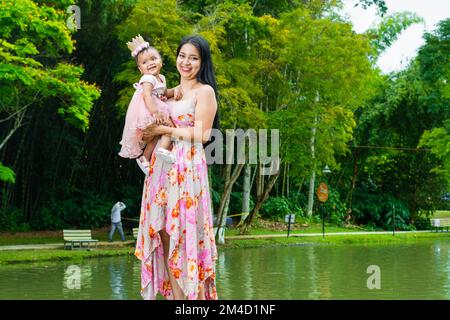 ritratto di una donna latina che tiene in braccio il suo bambino piccola brunetta mentre fa una passeggiata e cammina lungo il bordo di un lago in un parco. Foto Stock