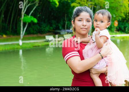 ritratto di una donna latina che tiene tra le braccia una piccola bruna, in piedi accanto ad un lago e circondata da verde natura Foto Stock