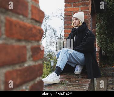 Una ragazza in abiti caldi e morbidi si siede nell'apertura di un muro di mattoni e guarda via Foto Stock