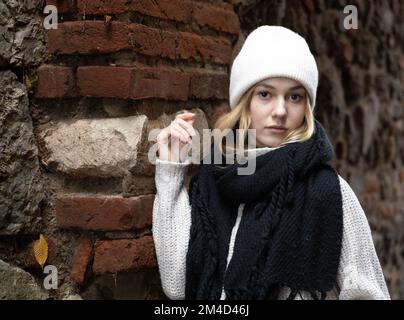 Una ragazza in abiti caldi e morbidi si trova vicino a un muro di mattoni e pietra testurizzati e guarda la macchina fotografica Foto Stock