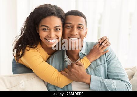 Felice giovane ragazza afro-americana sposando marito sul divano e guardando la macchina fotografica nel soggiorno interno Foto Stock