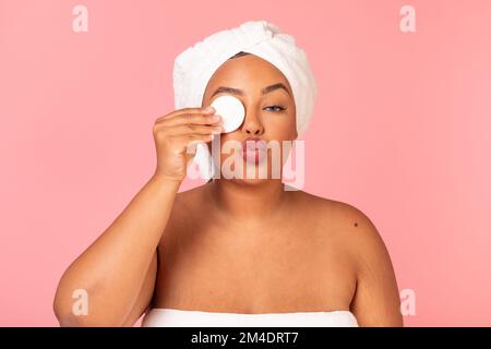 Bella afro americano corpo positivo donna rimuovere il trucco con tampone di cotone e fare aria bacio, sfondo rosa Foto Stock