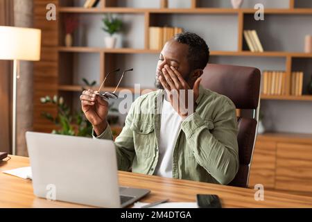 Stanco triste maschio nero maturo toglie i suoi occhiali, strofinando gli occhi, soffrendo di lavoro eccessivo e di lavoro eccessivo Foto Stock