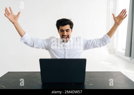 Studio ritratto di allegro eccitato lavoratore ufficio maschile urlare eureka alzando le mani, risolvendo duro lavoro seduto alla scrivania in camera con luce Foto Stock
