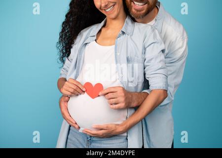 Giovane ragazzo sorridente europeo che abbraccia una donna incinta in un cuore casual che tiene sulla pancia grande Foto Stock