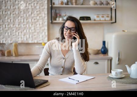 Ritratto di felice donna caucasica che lavora da casa, parlando sul cellulare e utilizzando il computer portatile, seduto in cucina Foto Stock