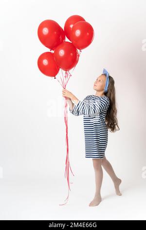 Nosy ragazza che tiene mazzo di palloncini rossi in due mani e guardando palloncini. Modello giocoso di dieci anni con capelli lunghi in abito a righe blu bianco Foto Stock