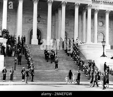 I portatori del corpo che trasportano la scrigno del presidente Kennedy sui gradini centrali del Campidoglio degli Stati Uniti, seguiti da una guardia di colore che tiene la bandiera del presidente degli Stati Uniti. Sua moglie Jacqueline Kennedy e i suoi figli, Caroline Kennedy e John F. Kennedy Jr. Stanno per iniziare i gradini, il 24 novembre 1963. Foto Stock