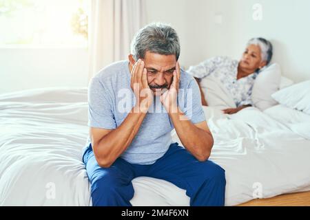 A volte non siamo d'accordo sulle cose. un uomo maturo irritato seduto su un letto con sua moglie sdraiata sullo sfondo dopo aver avuto un argomento a casa. Foto Stock