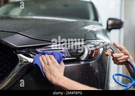 Primo piano immagine ritagliata di mano di giovane lavoratore di auto servizio, strofinando e lucidando i fari e il cofano di moderna auto grigia con panno in microfibra blu sotto acqua ad alta pressione. Foto Stock