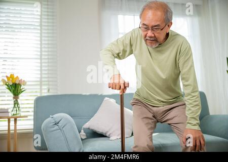 Asian Old Man con occhiali che battono per alzarsi dal divano con bastone da passeggio Foto Stock