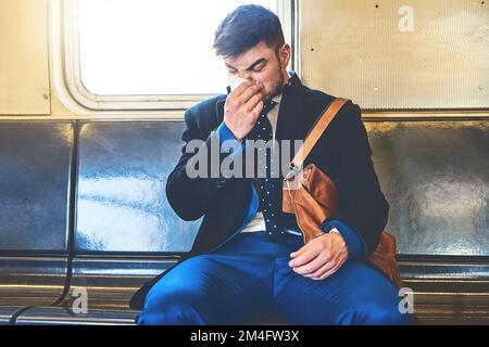 Meglio non ammalarmi. un giovane che guarda irritato che soffia il naso mentre è seduto all'interno di un treno per arrivare al lavoro. Foto Stock