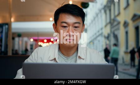 Focalizzato asiatico uomo turista seduto all'aperto navigare in rete sociale sul computer portatile utilizzando wi-fi gratuito millennial di viaggio blogger maschile digitando sul computer Foto Stock