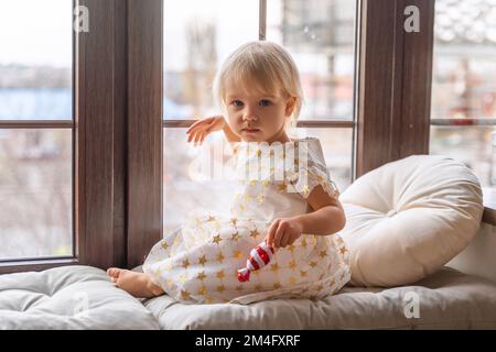 Ragazza di 2 anni siede sul davanzale. Una bambina bionda in un vestito bianco sul davanzale guarda la macchina fotografica e tiene una caramella in mano. Foto Stock