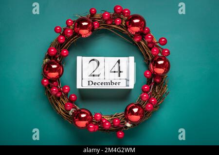 Corona di Natale decorata con bacche rosse, calendario in legno data 24 dicembre isolato su sfondo verde concetto di preparazione di Natale, atmosfera auguri carta fatto a mano corona di Natale piatto laici. Foto Stock