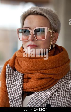 Donna ritratto capelli biondi corti in caldo maglia sciarpa ocra, giacca bipetto e occhiali alla moda. Moda autunno o primavera, femmina f Foto Stock