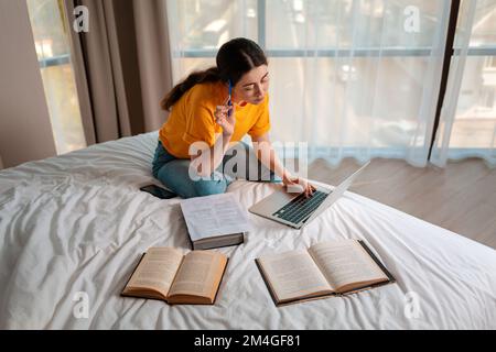 Formazione online. Una donna in abiti casual è seduta su un letto e al lavoro su un computer portatile. I libri si trovano l'uno accanto all'altro. Il concetto di cou online Foto Stock