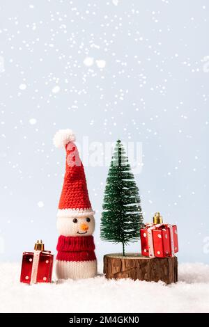 La fiaba di Natale composizione di un pupazzo di neve lavorato a maglia in un cappello e un maglione di Babbo Natale. Piccolo albero di Natale con due regali rossi Foto Stock