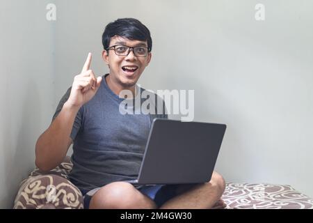 Ritratto felice occhio asiatico occhiali uomo è puntando dito indice e guardando il notebook su una camera da letto per il lavoro da casa trovare nuove idee concetto con natura Foto Stock