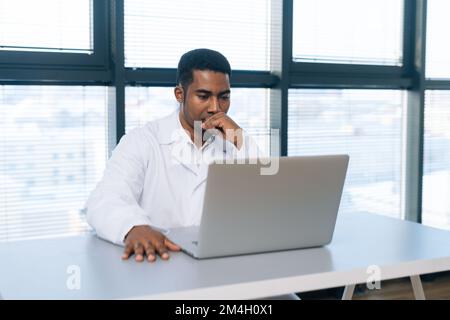 Ritratto del giovane afro-americano medico professionista maschio pensivo in bianco uniforme pensando guardando lo schermo del computer portatile seduto alla scrivania. Foto Stock
