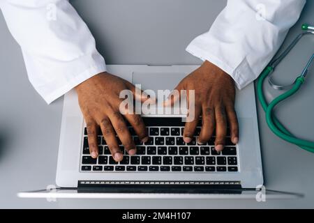 Primo piano vista ad angolo alto di un medico maschio nero irriconoscibile professionista in uniforme bianca di lavoro digitando sul computer portatile seduto alla scrivania con Foto Stock