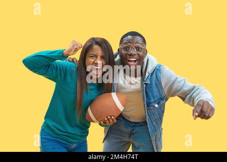 Coppia di diversità etnica eccitata godendo il calcio americano insieme isolato su sfondo giallo Foto Stock